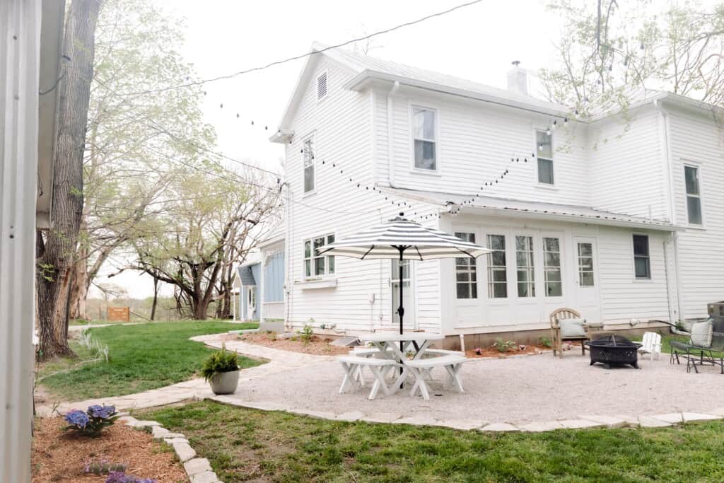 the back of the 1800's Victorian farmhouse with a gravel patio with table and umbrella and beautiful landscaping
