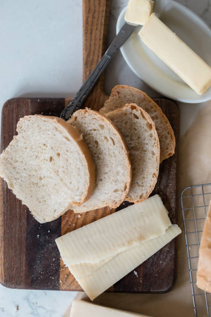 overhead photo of slices of sourdough sandwich bread on a wood cutting board with slice cheese and a plate of butter to the back right