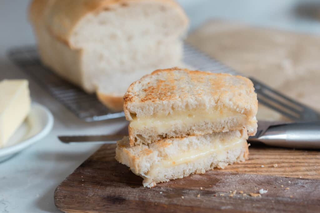 a sourdough grilled cheese sliced in half and stacked on top of each other on a wood cutting board with a sourdough loaf in the background