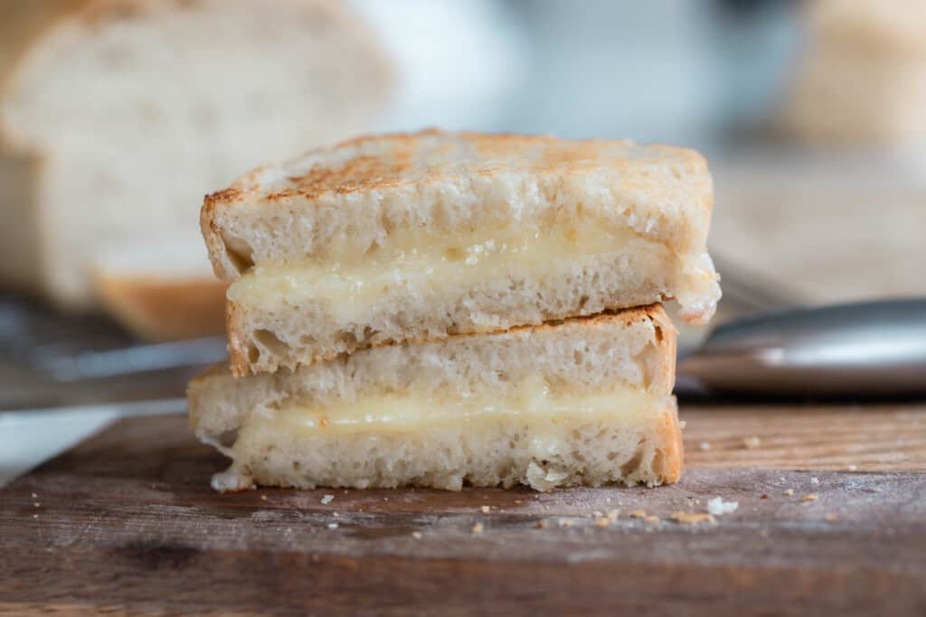 close up of two slices of sourdough grilled cheese stacked on a wood cutting board with knife and loaf of bread in the back