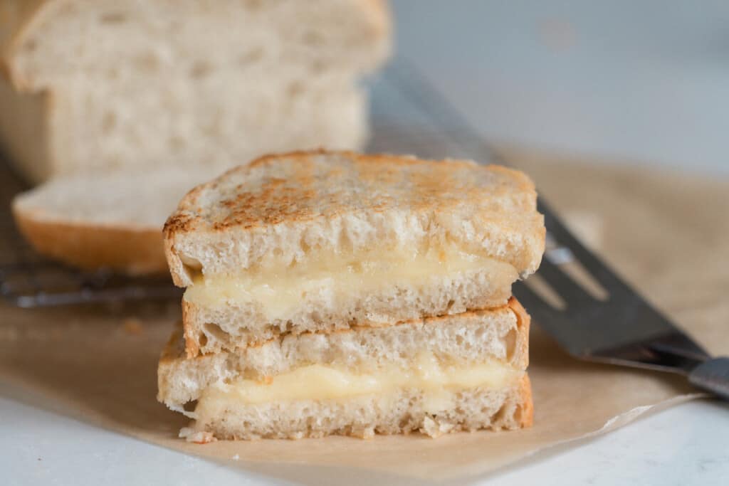 two halves of grilled cheese stacked on top of each other on parchment paper with a metal spatula to the right and a loaf of bread in the background