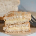 two halves of grilled cheese stacked on top of each other on parchment paper with a metal spatula to the right and a loaf of bread in the background