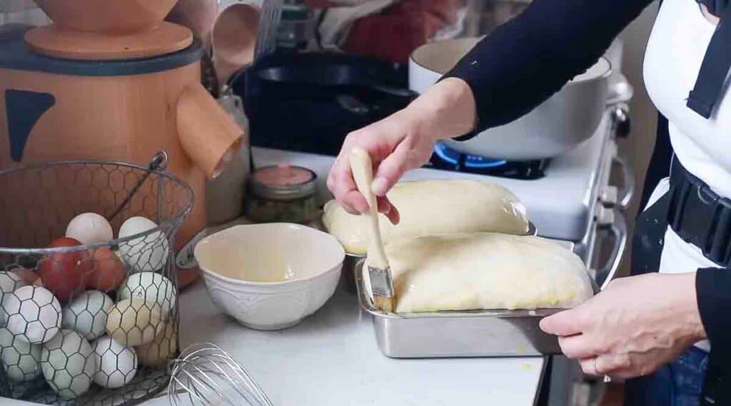 woman using a pastry brush to brush on egg wash onto a loaf of sourdough bread dough in a loaf pan