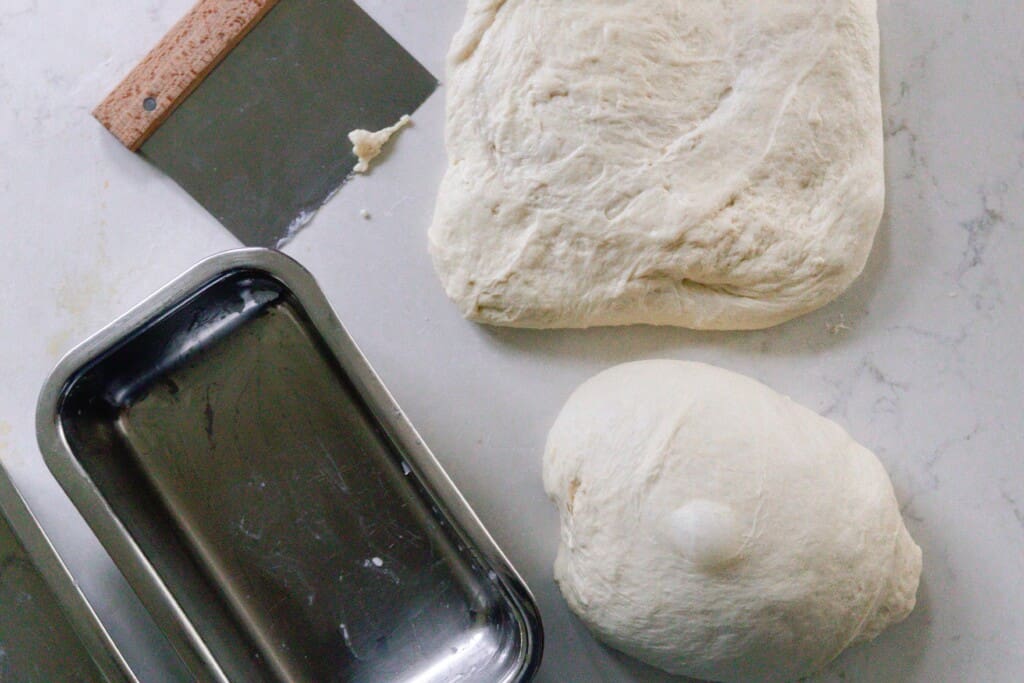 two pieces dough on a white countertop. One is in a rectangle and the other in a ball next to a bench scraper and a loaf pan