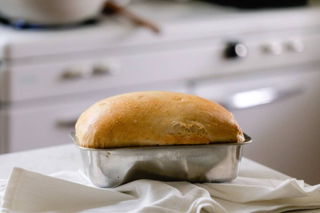 loaf of sourdough sandwich bread in a stainless loaf pan on a white countertop with a white towel in the front