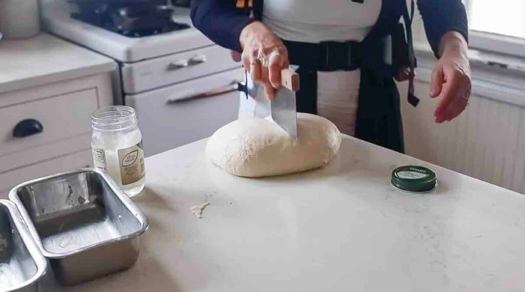 woman using a bench scraper to slice sourdough bread dough in half on a white countertop