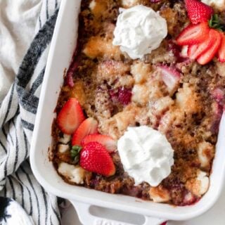 sourdough strawberry cobbler topped with fresh whipped cream and sliced strawberries. The cobbler is in a white baking dish on a white and black stripped towel