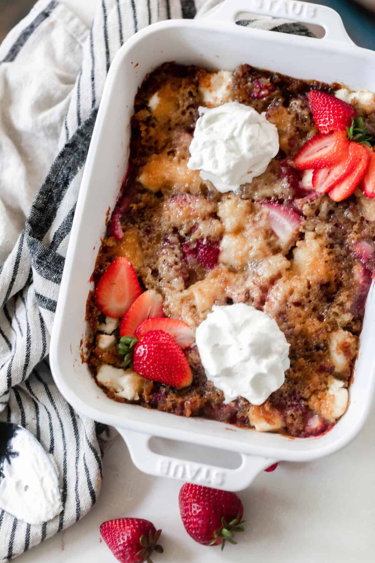 sourdough strawberry cobbler topped with fresh whipped cream and sliced strawberries. The cobbler is in a white baking dish on a white and black stripped towel