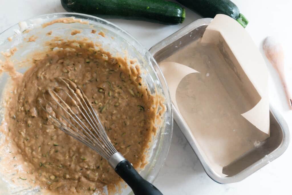 zucchini bread batter in a glass bowl with a parchment lined stainless steel loaf pan to the right