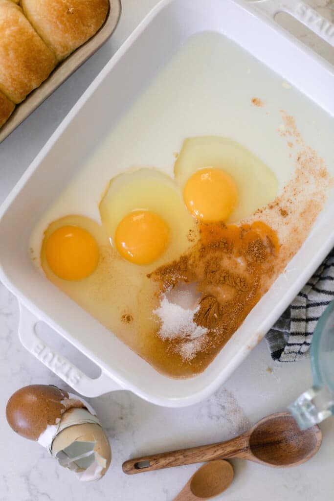 eggs sugar, and cinnamon in a shallow baking ban on a white countertop with measuring spoons to the front right