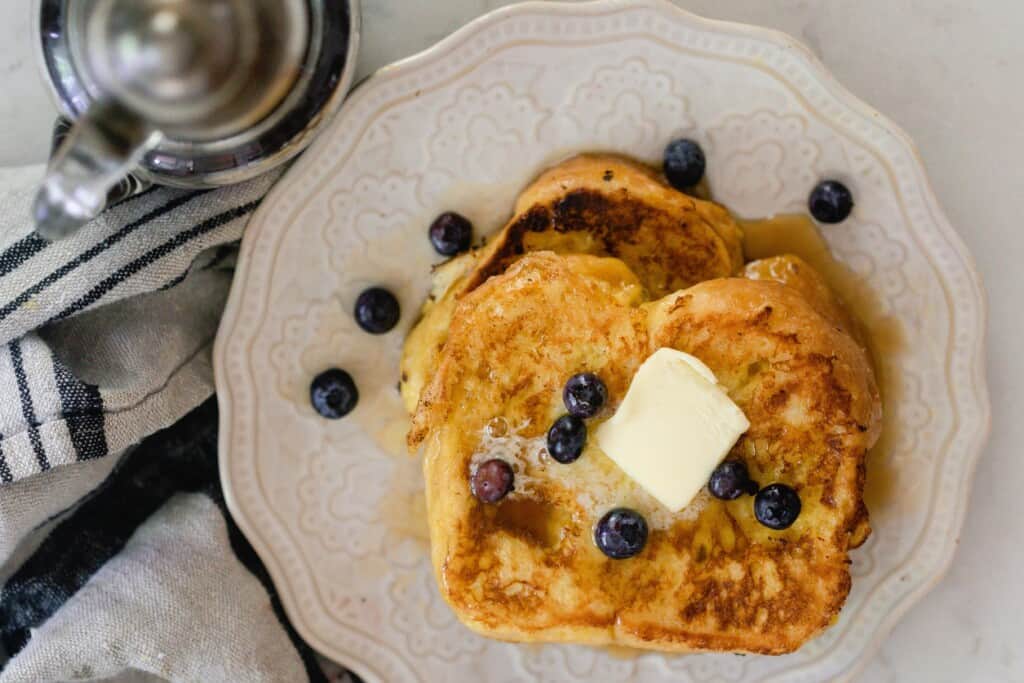 two brioche French toast slices stack on top of each other and topped with butter and fresh blueberries on a cream colored place on a stripped napkin