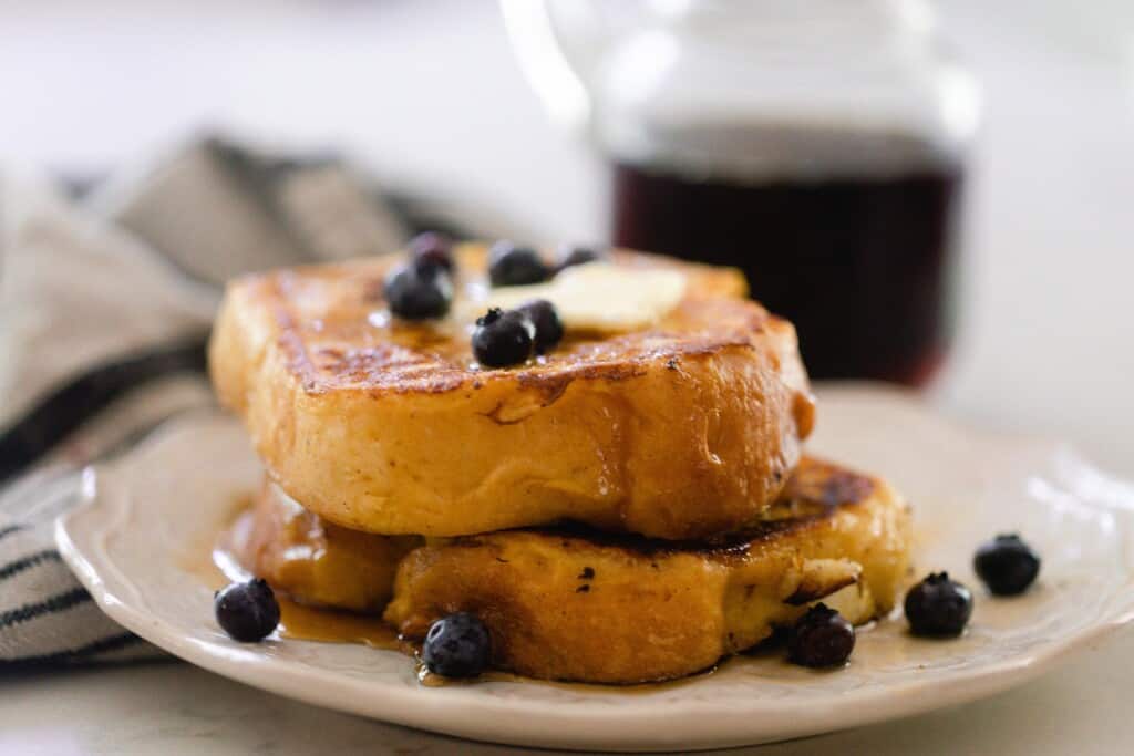 side view of thick sliced brioche French toast topped with a. pat of butter and blueberries on a cream colored plate with a jar of maple syrup in the background.