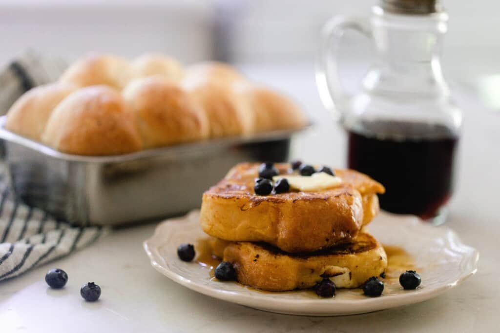 two thick slices of brioche French toast topped with butter and fresh blueberries on a white plate with a small pitcher of maple syrup and a loaf of brioche in a loaf pan placed on a black and white stripped towel.