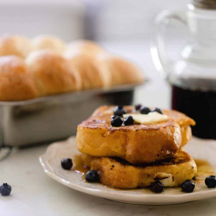 two thick slices of brioche French toast topped with butter and fresh blueberries on a white plate with a small pitcher of maple syrup and a loaf of brioche in a loaf pan placed on a black and white stripped towel.