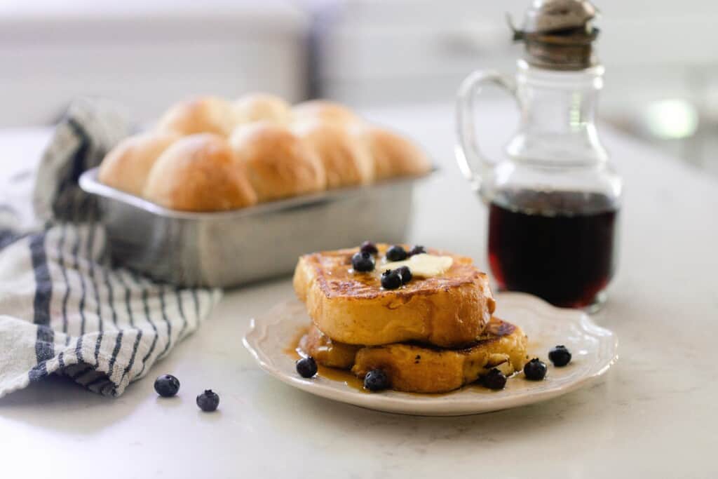 brioche French toast stacked on a cream colored plate and topped with fresh blueberries and butter.  A small pitcher of maple syrup and a loaf of brioche in a loaf pan are behind the plate