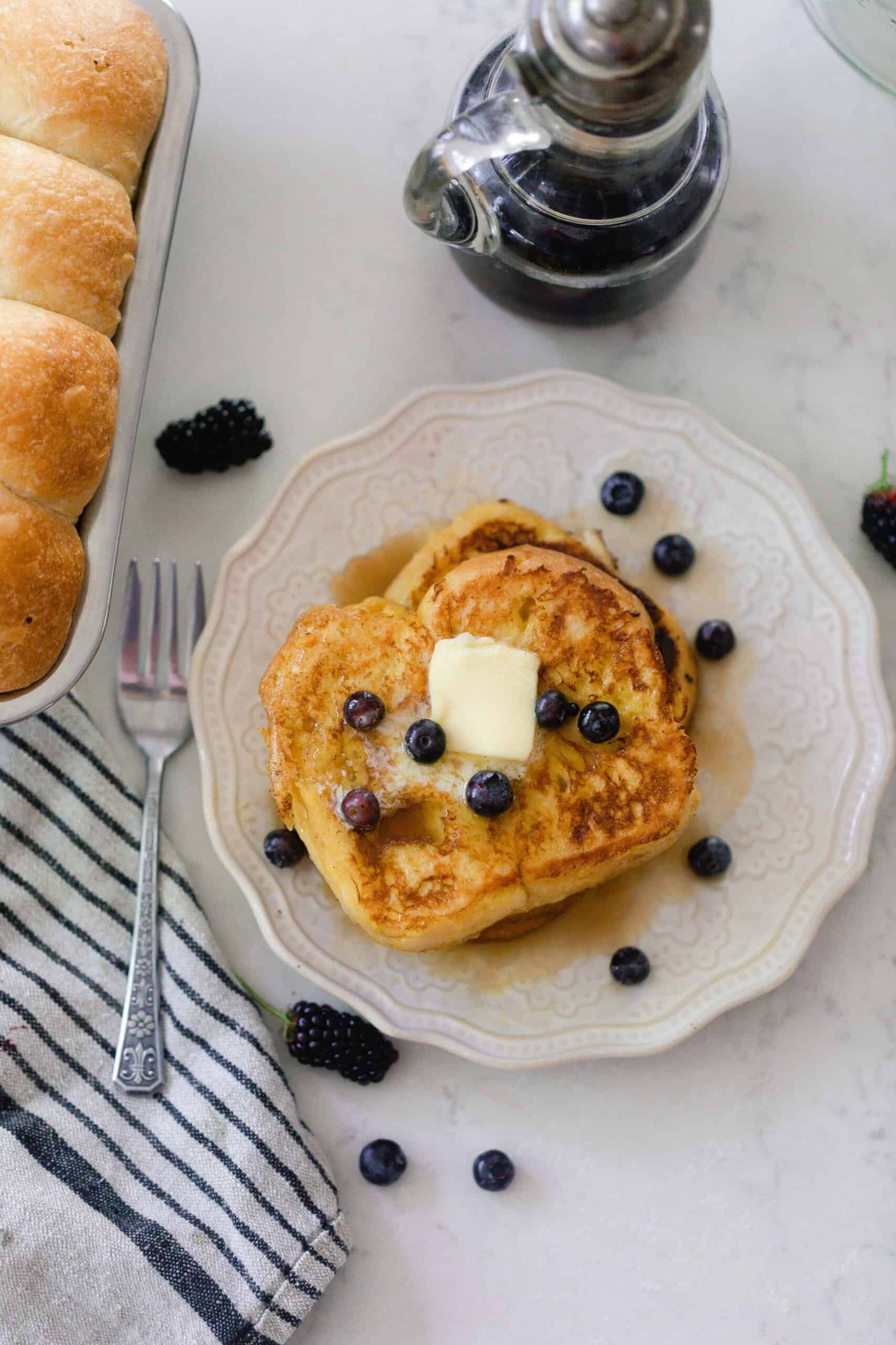 overhead picture of two brioche French toast slices stacked on top pf each other and topped with butter and fresh blueberries on a cream colored plate. The plate is on a white quartz countertop with a stripped towel and a loaf go brioche to the left