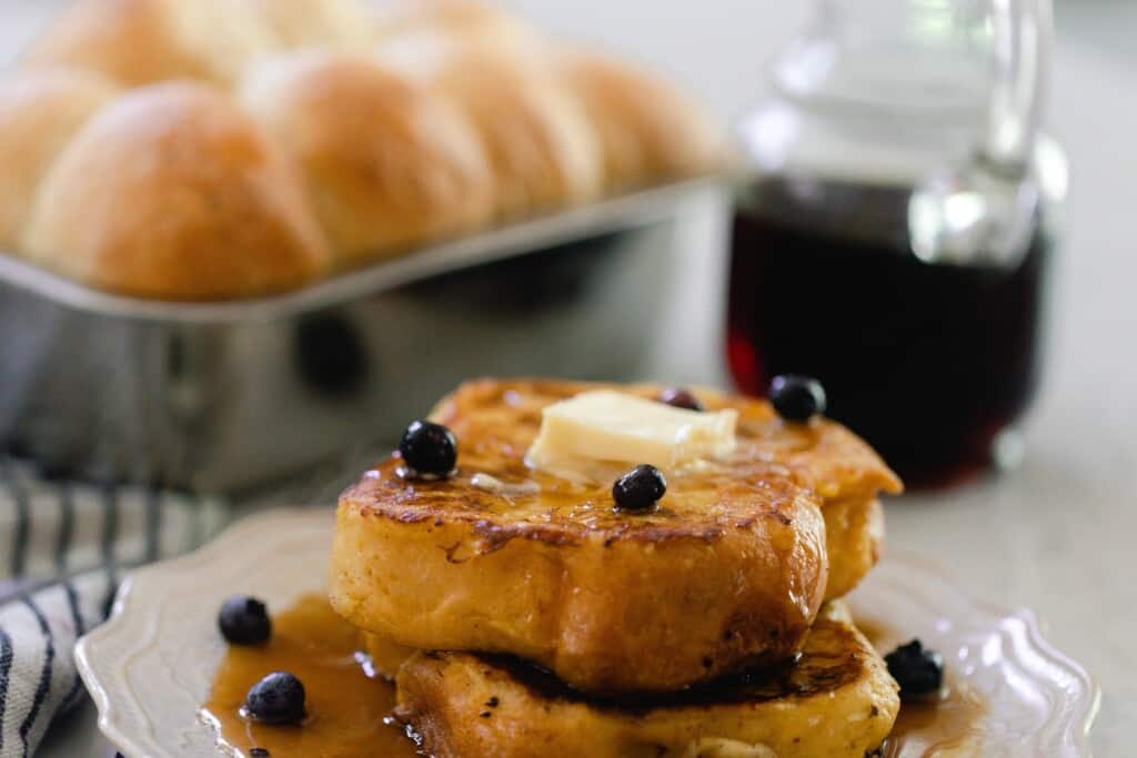 close up picture of two thick slices of brioche French toast topped with butter and blueberries on a white plate with a small picture of syrup and a loaf of brioche in a loaf pan in the background