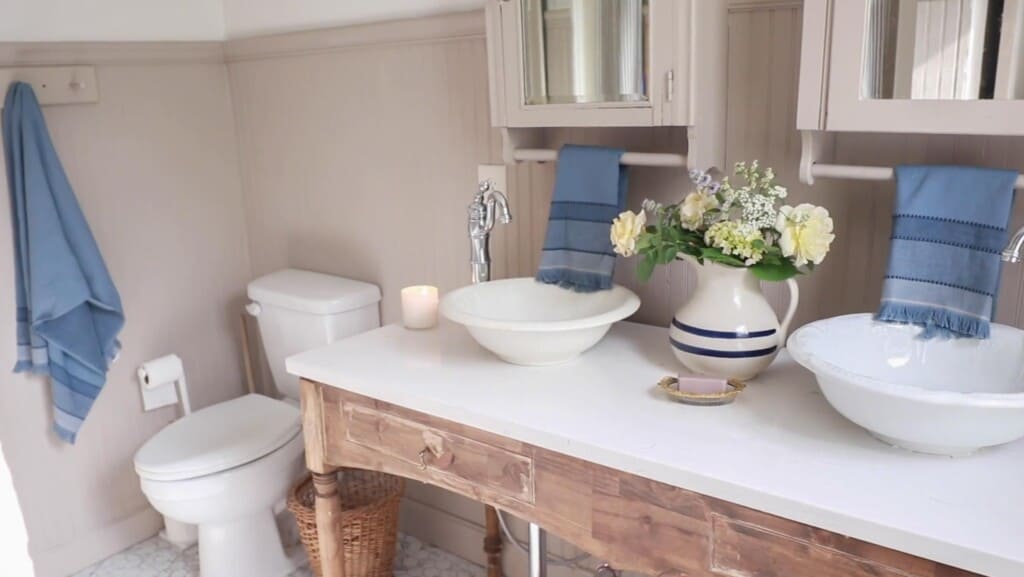 farmhouse kids bathroom with a custom antique inspired vanity with a marble top and antique bowl vessel sinks. Two medicine cabinets hang above the sinks with blue towels hanging off the cabinets