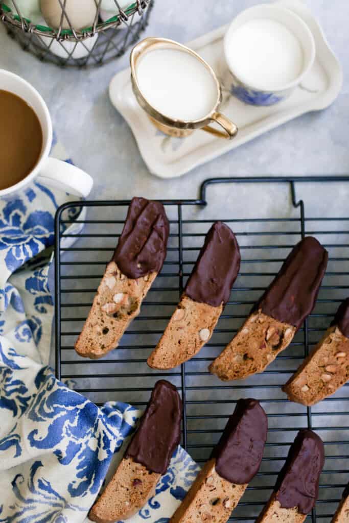sourdough almond biscotti dipped in chocolate and drying on a wire rack. Two white serving cups with cream and sugar and a cup of coffee are next to the baking rack