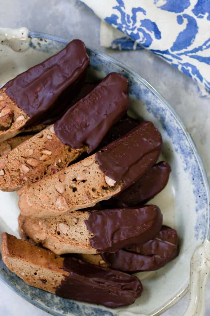 sourdough biscotti dipped in chocolate layered on a white and blue platter  with a blue and white floral towel in the top right corner