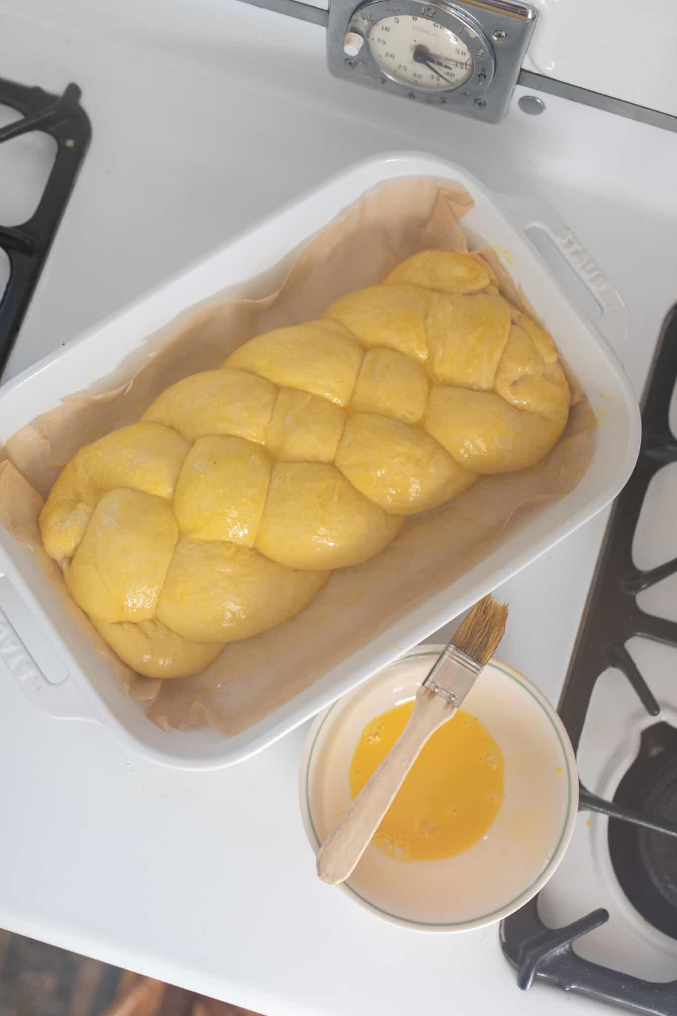 sourdough challah dough braided and topped with an egg wash resting in a white baking dish on a antique stove