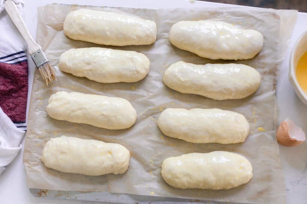 sourdough hot dog bun dough shaped onto parchment paper, allowed to rise a final time, and an egg wash brushed over