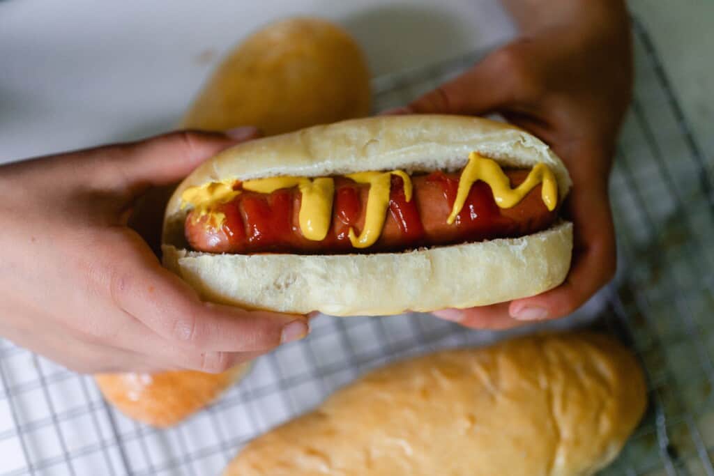 hands holding a hot dog with ketchup and mustard drizzled on top in a sourdough hot dog bun.