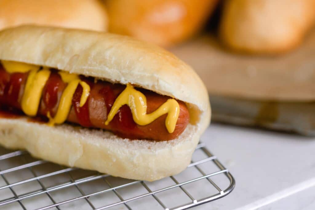 hot dog with ketchup and mustard in a homemade sourdough hot dog bun on a wire rack. More hot dog buns in the background