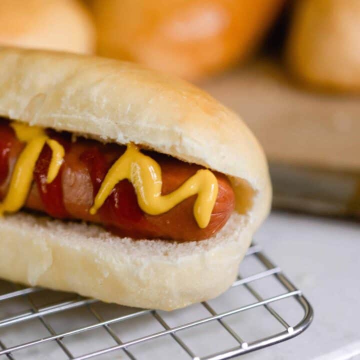 hot dog with ketchup and mustard in a homemade sourdough hot dog bun on a wire rack. More hot dog buns in the background