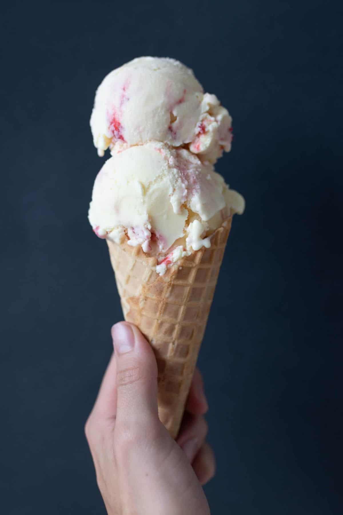 A hand holding an ice cream cone with two scoops of strawberry cheesecake ice cream.