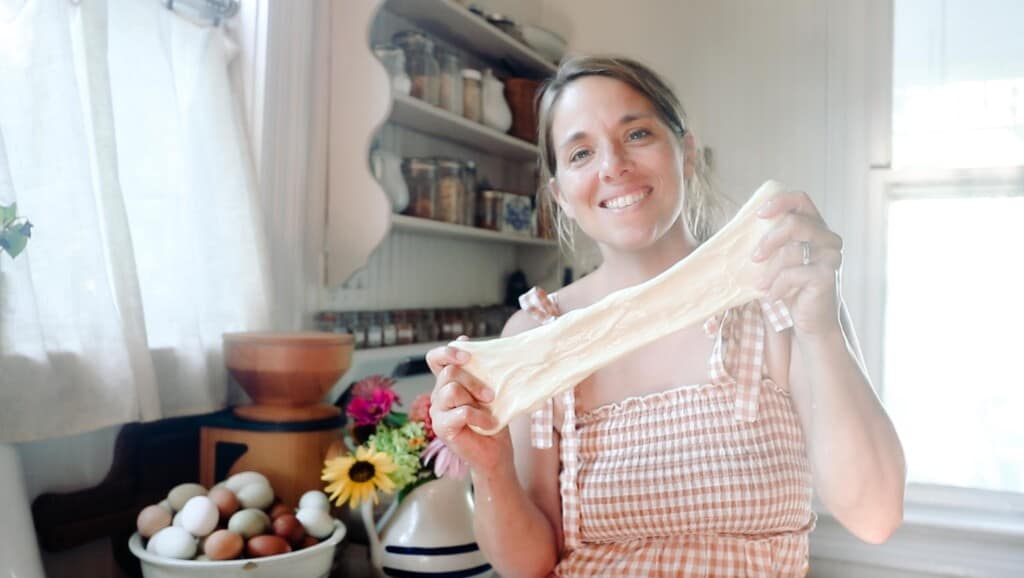 woman wearing a pink plaid dressing stretching homemade mozzarella cheese between her hands in her kitchen