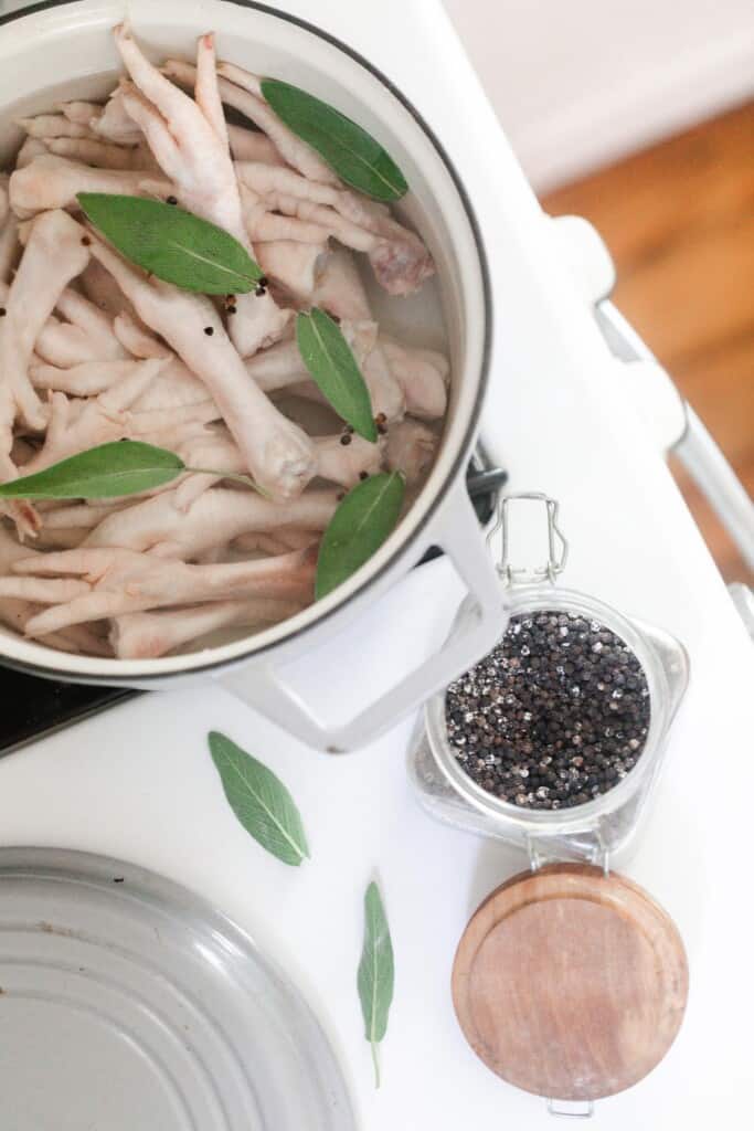 white dutch oven filled with chicken feet, water, and safe leaves with salt and peppercorns in jars to the right