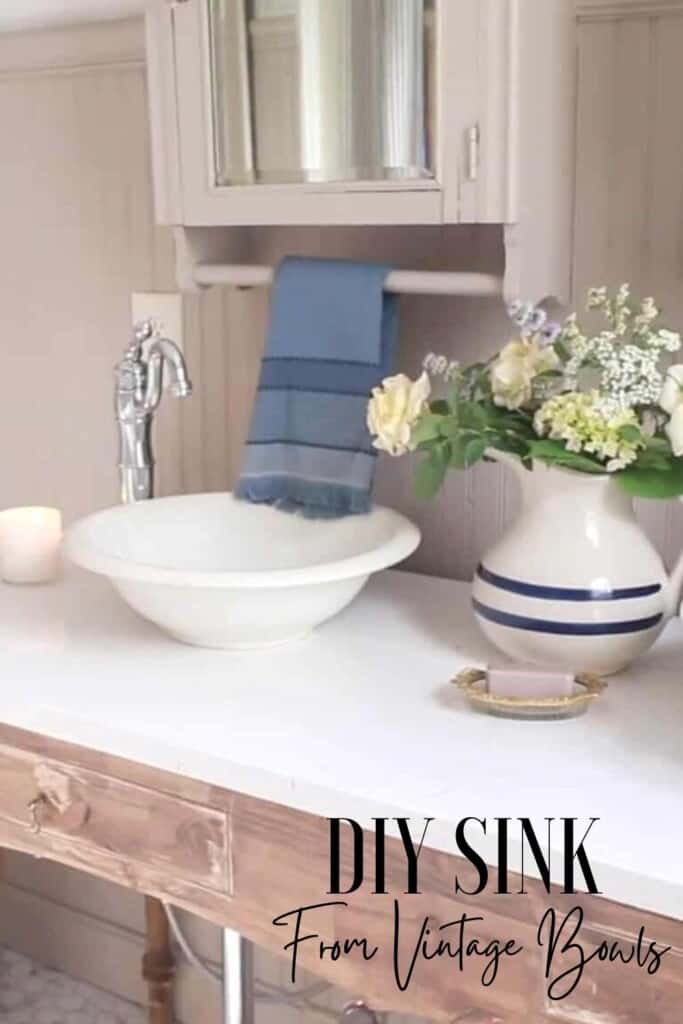 DIY sink made from a vintage bowl sits on top of a marble countertop and handmade vanity. A pitcher with flowers is to the right and a antique medicine cabinet with blue towel hanging from the bar is directly above the sink