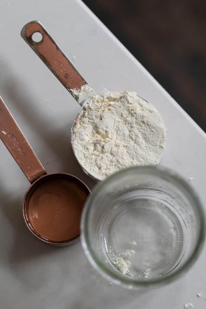 a jar of water and two measuring cups with einkorn flour and water on a white countertop