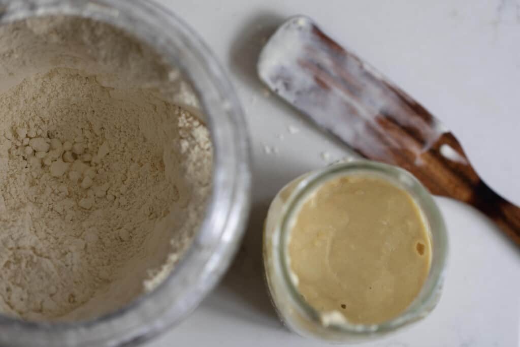 two jars; one with einkorn flour and another smaller jar with einkorn sourdough starter with a wood stirrer.