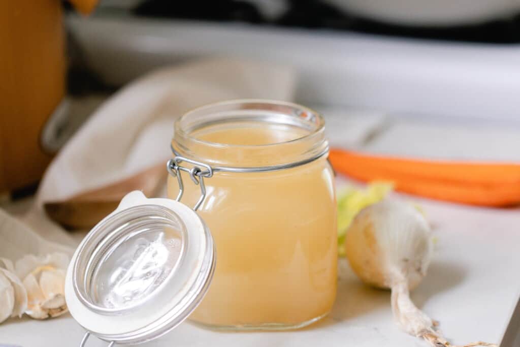 jar with swing top lid filled with chicken broth on a white countertop with onions and garlic surrounding the jar