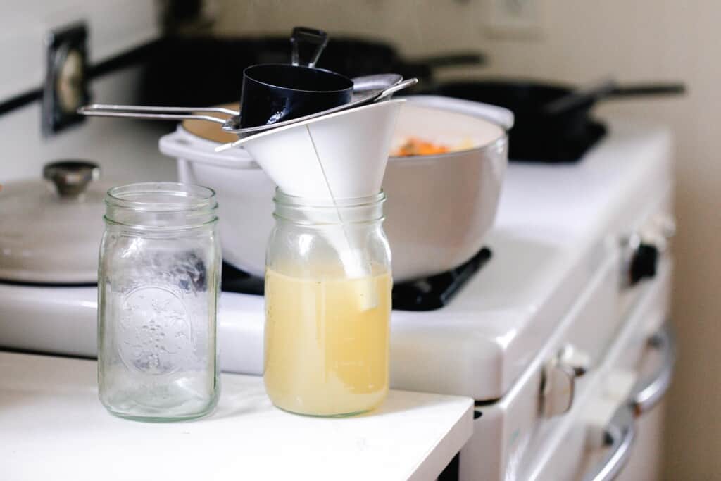 straining bone broth into mason jars with a white funnel and fine mesh strainer on a white countertop