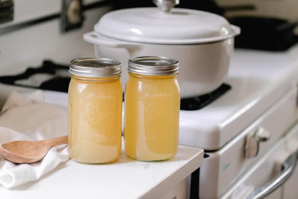 two jars of homemade bone broth in mason jars on a white quartz countertop with a gray dutch oven in the background