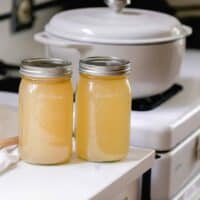 two jars of homemade bone broth in mason jars on a white quartz countertop with a gray dutch oven in the background