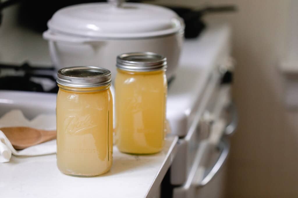 two jars of homemade chicken bone broth on a white countertop with a white stove with a dutch oven in the background