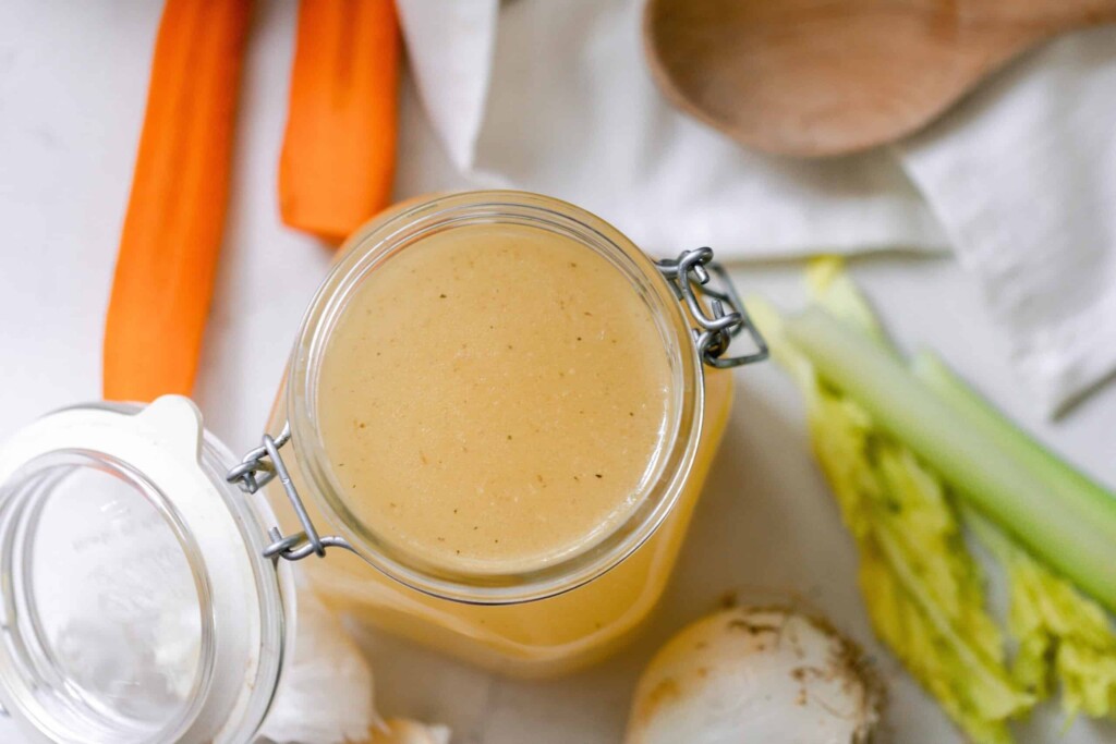 overhead photo of a chicken broth in a swing top jar with celery, garlic, carrots and a wooden spoon surrounding the jar