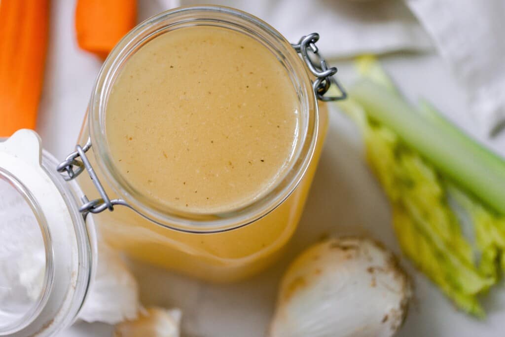 overhead photo of bone broth in a swing top jar. The jar is surrounded by carrots, celery, and onions.