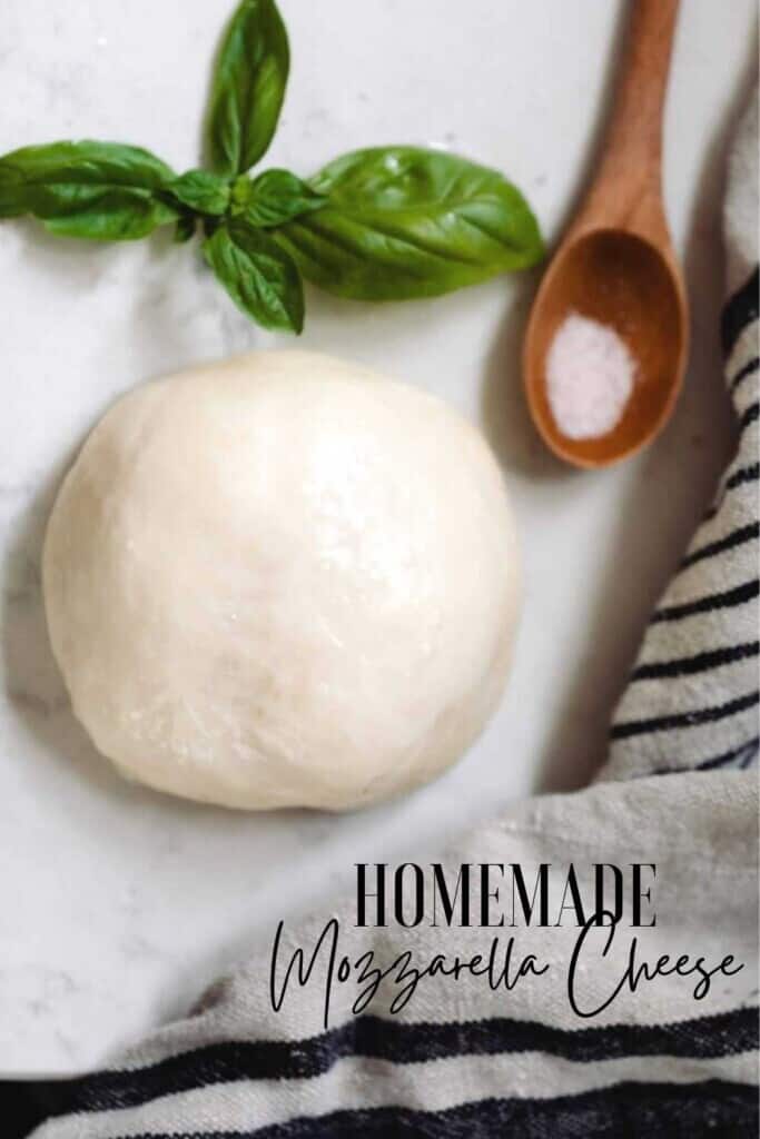 ball of homemade mozzarella cheese on a white countertop with a fresh sprig of basil, a wooden spoon with salt, and a black and white stripped towel surrounding the cheese