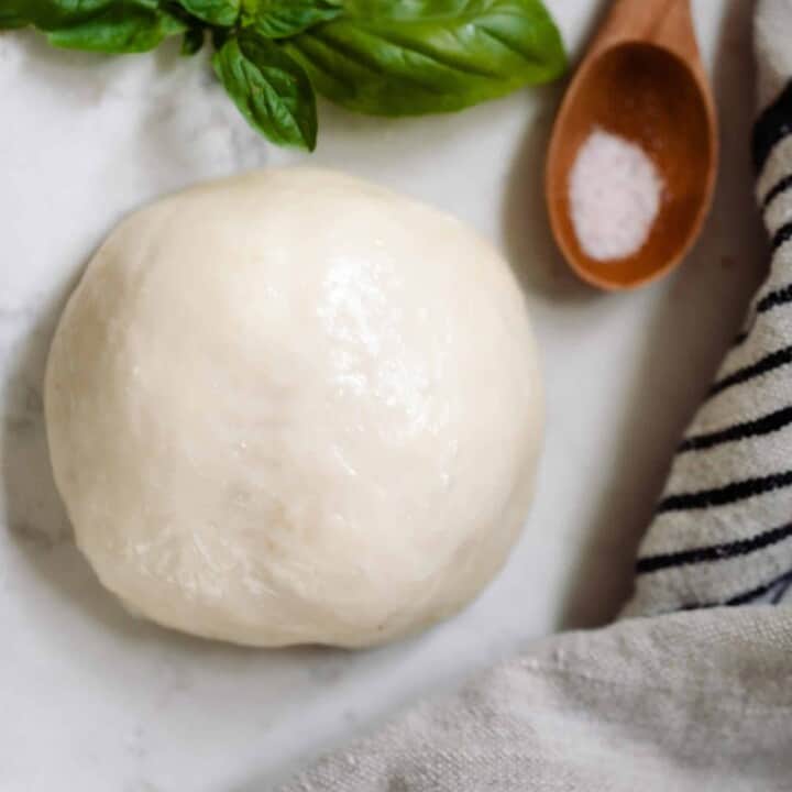 ball of homemade mozzarella cheese on a white countertop with a fresh sprig of basil, a wooden spoon with salt, and a black and white stripped towel surrounding the cheese