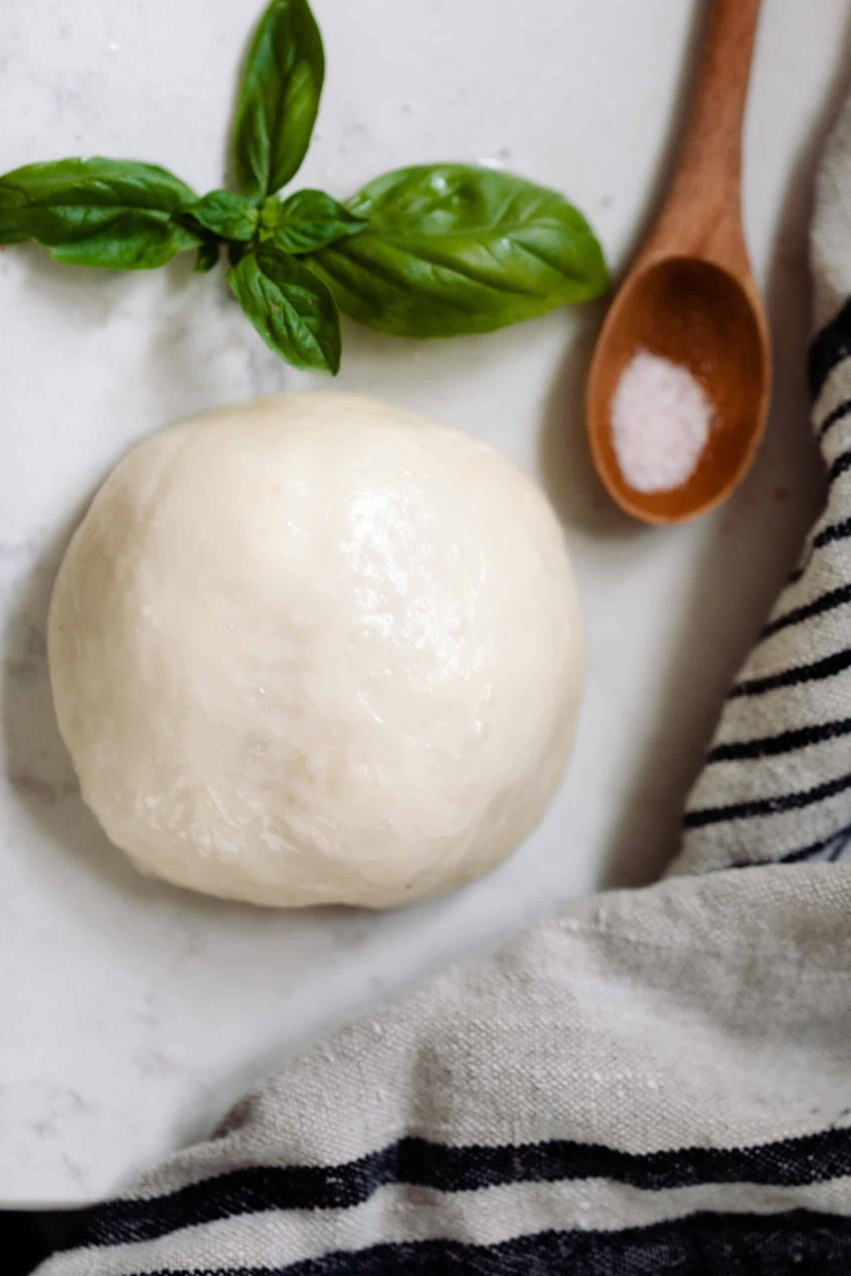 ball of homemade mozzarella cheese on a white countertop with a fresh sprig of basil, a wooden spoon with salt, and a black and white stripped towel surrounding the cheese