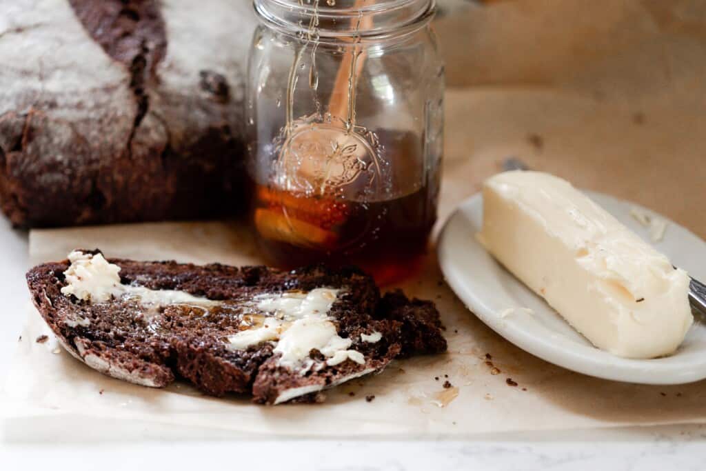 slice of chocolate sourdough bread topped with butter and honey. A small white dish with butter, and a jar of honey are in the background