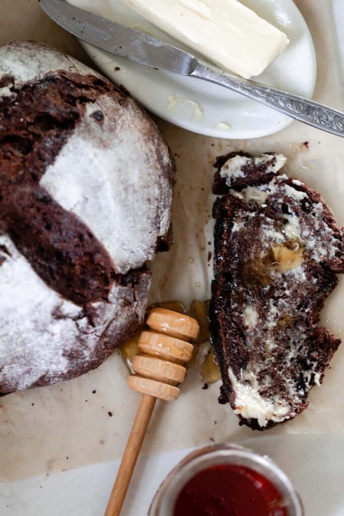 overhead picture of a loaf of sourdough chocolate bread with a slice of the bread to the right by honey and butter