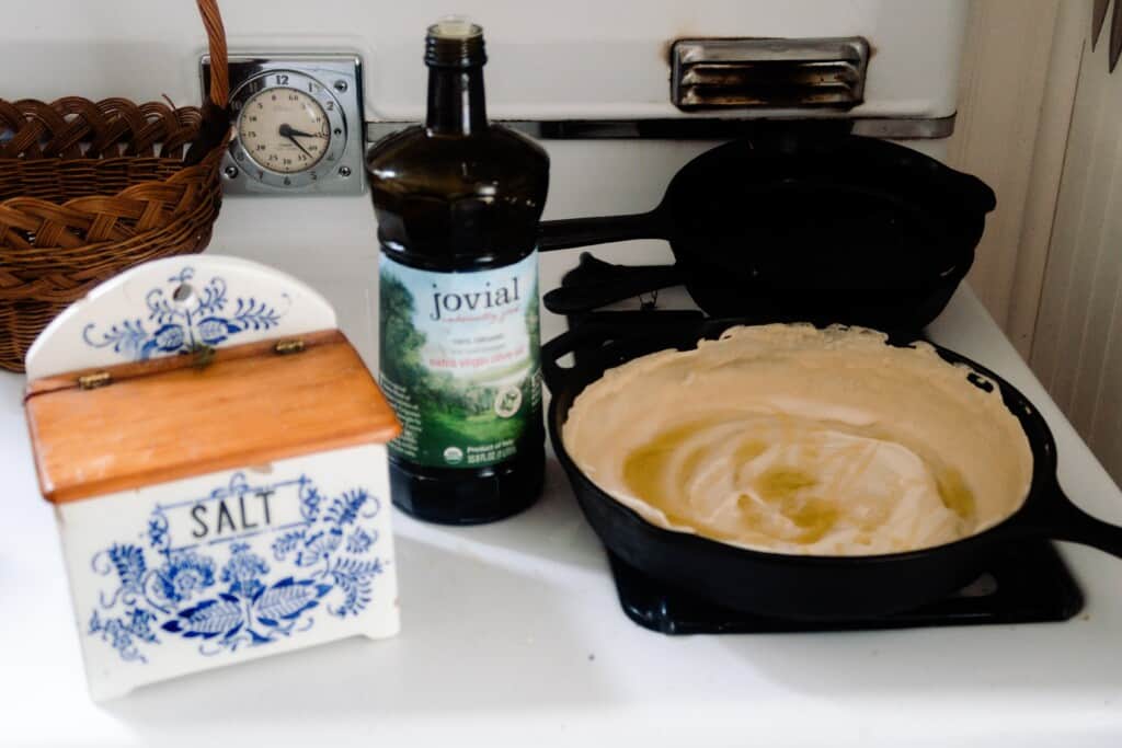 sourdough starter cooking in a cast iron skillet topped with olive oil. The skillet in on a white vintage stove with a bottle of olive oil and a vintage container of salt.