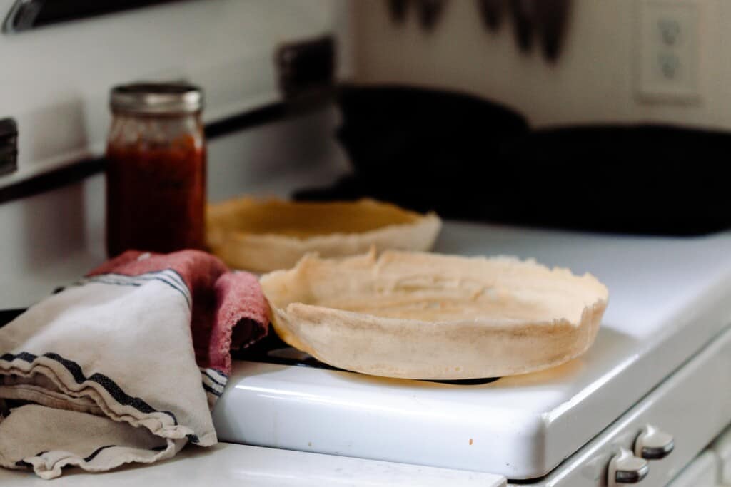 two sourdough discard pizza crusts on a white vintage stove with a a cream, black, and red towel to the left next to a jar of sauce.