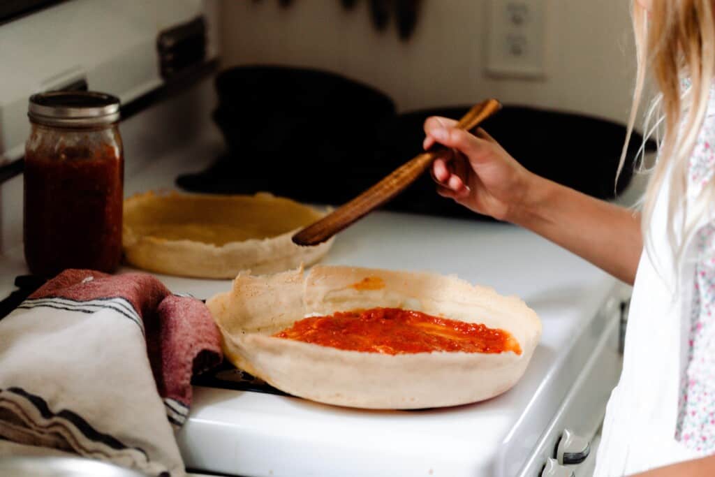 a girl with blonde hair spreading pizza sauce on a sourdough pizza crust that is on a white vintage stove